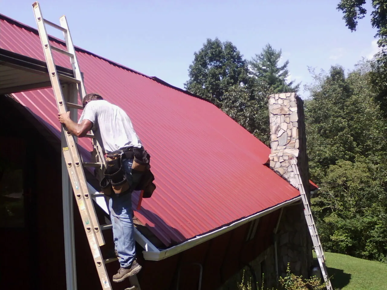Roofer on a ladder beside a red metal roof and stone chimney — Soffit & Fascia Repair by Roofing Brothers in Lostine